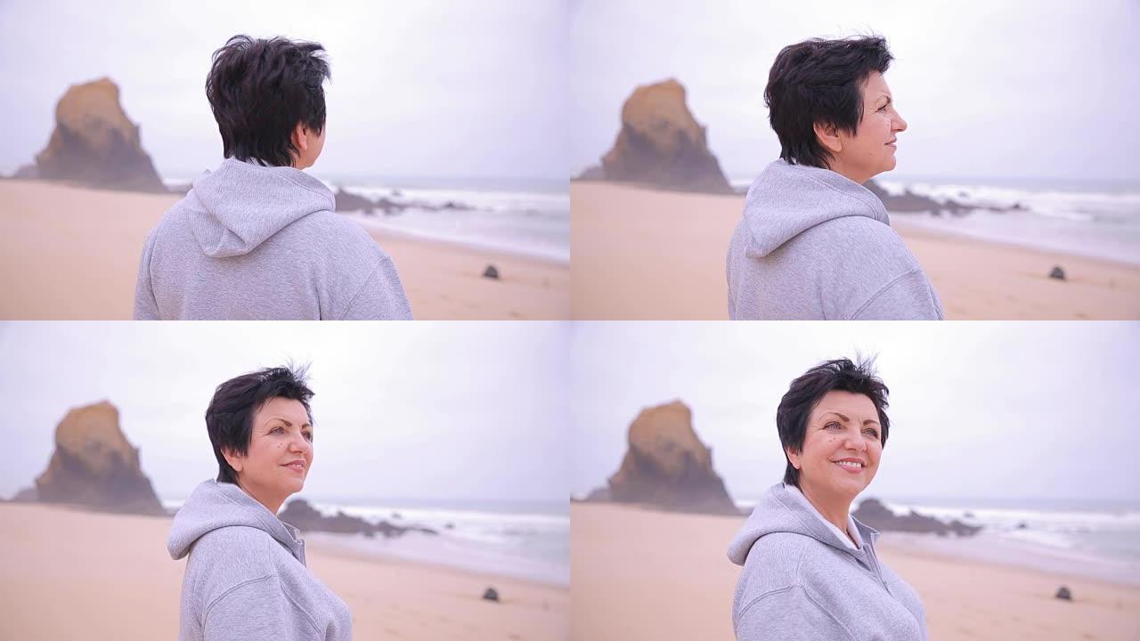 迷人的活跃的高级女性在荒芜的海滩上欣赏海洋，对着镜头微笑。