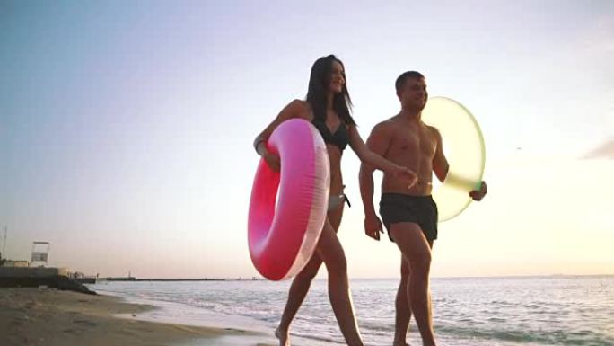 幸福的年轻夫妇在日落时带着橡胶圈在沙滩上散步，慢动作