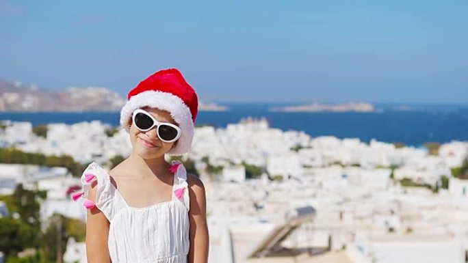 米克诺斯户外背景的白色连衣裙和红色帽子迷人女孩。孩子在希腊米科诺斯岛上典型的希腊传统村庄的街道上，白