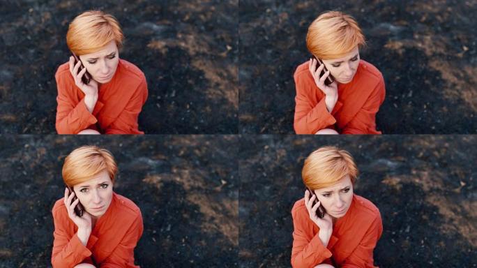 非常悲伤的红发女人坐在烧焦的草地上，拿着电话放在耳朵上。概念: 需要帮助、悲伤、孤独
