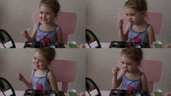 金发三岁的婴儿在她的脸上画彩色条纹看着镜子坐在桌子旁