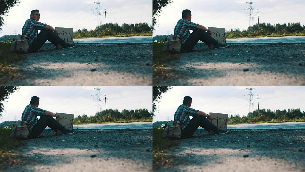 男子坐在乡村的路上，用纸板标牌城市。搭便车。等待