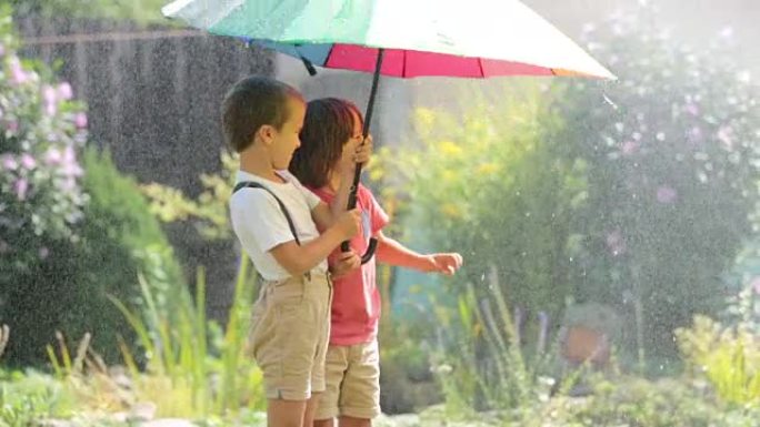 两个可爱的孩子，男孩兄弟，在后院洒水下玩着五颜六色的雨伞