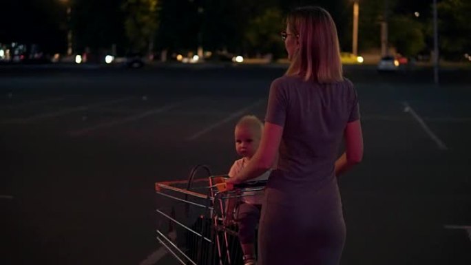 小婴儿坐在杂货店的手推车上，而她的母亲在购物后寻找汽车后，将手推车向前推，在超市旁边的停车场走