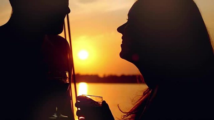 恋爱中的年轻幸福夫妇的剪影在游艇上接吻，并在惊人的日落中以慢动作喝杯葡萄酒。