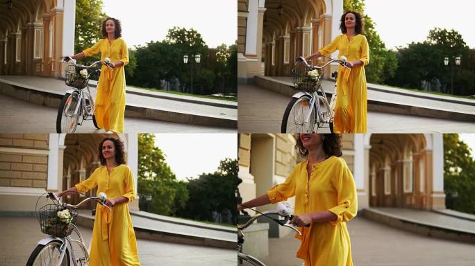 清晨，穿着黄色长裙的微笑的年轻女孩在城市的篮子里拿着鲜花，拿着城市自行车的车把。快乐的女人享受她的假