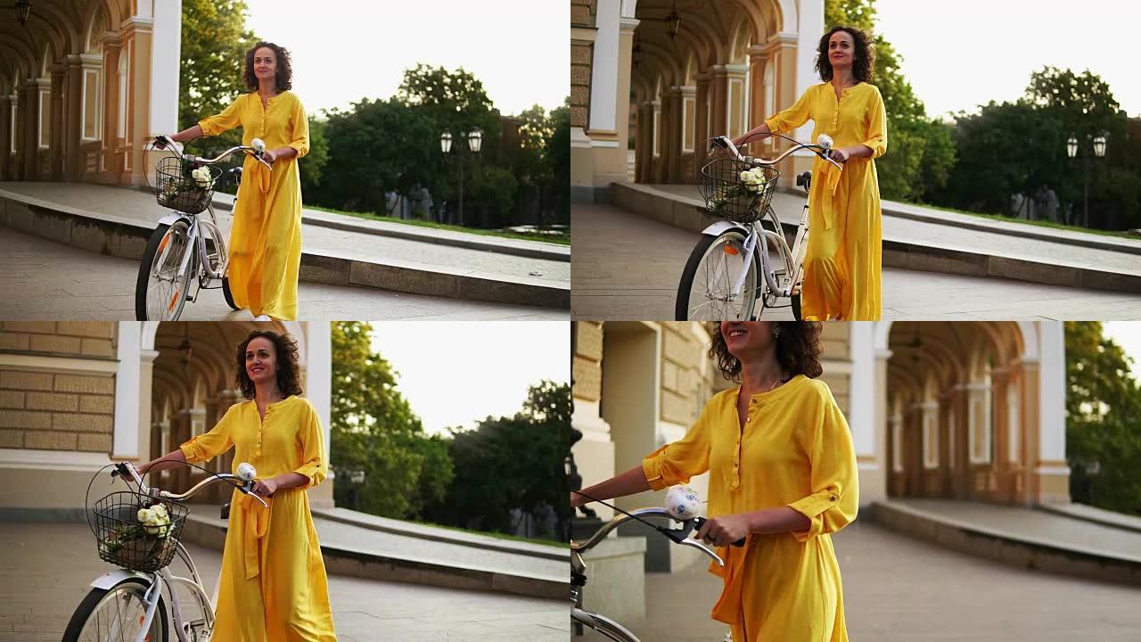 清晨，穿着黄色长裙的微笑的年轻女孩在城市的篮子里拿着鲜花，拿着城市自行车的车把。快乐的女人享受她的假