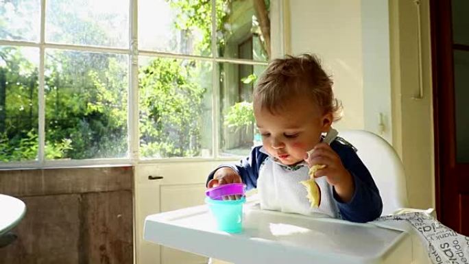 高脚椅上可爱的婴儿蹒跚学步的金发男孩的特写。婴儿吃蛋糕和玩婴儿玩具的肖像