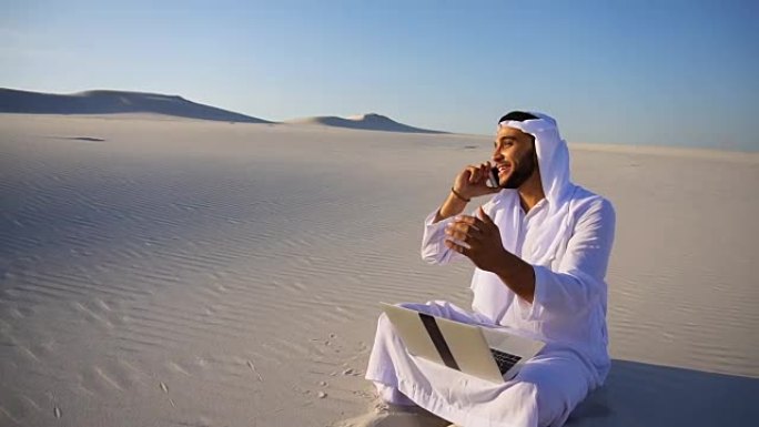 在夏天的大沙漠里，英俊的阿拉伯阿拉伯酋长男商人叫商业伙伴坐在沙地上，带着笔记本电脑