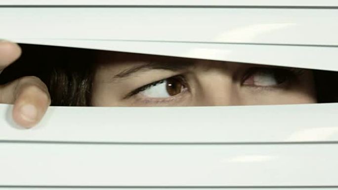 威尼斯百叶窗被年轻的棕色眼睛的女人拉开，她环顾四周，怀疑和恐惧，然后看着相机，感到非常恐惧和恐慌。