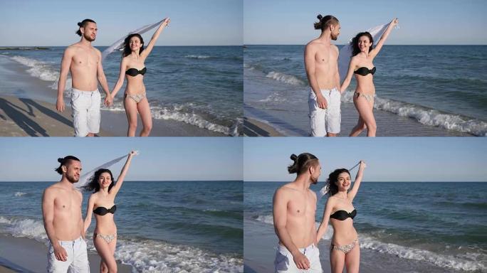 浪漫的散步，男人在海滩上手牵着女孩，朋友赤脚在沙滩上散步，风长出女人的头发和白布