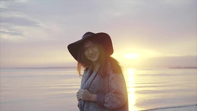 日落时分，在海边，戴着黑帽子和眼镜的年轻漂亮微笑的女人