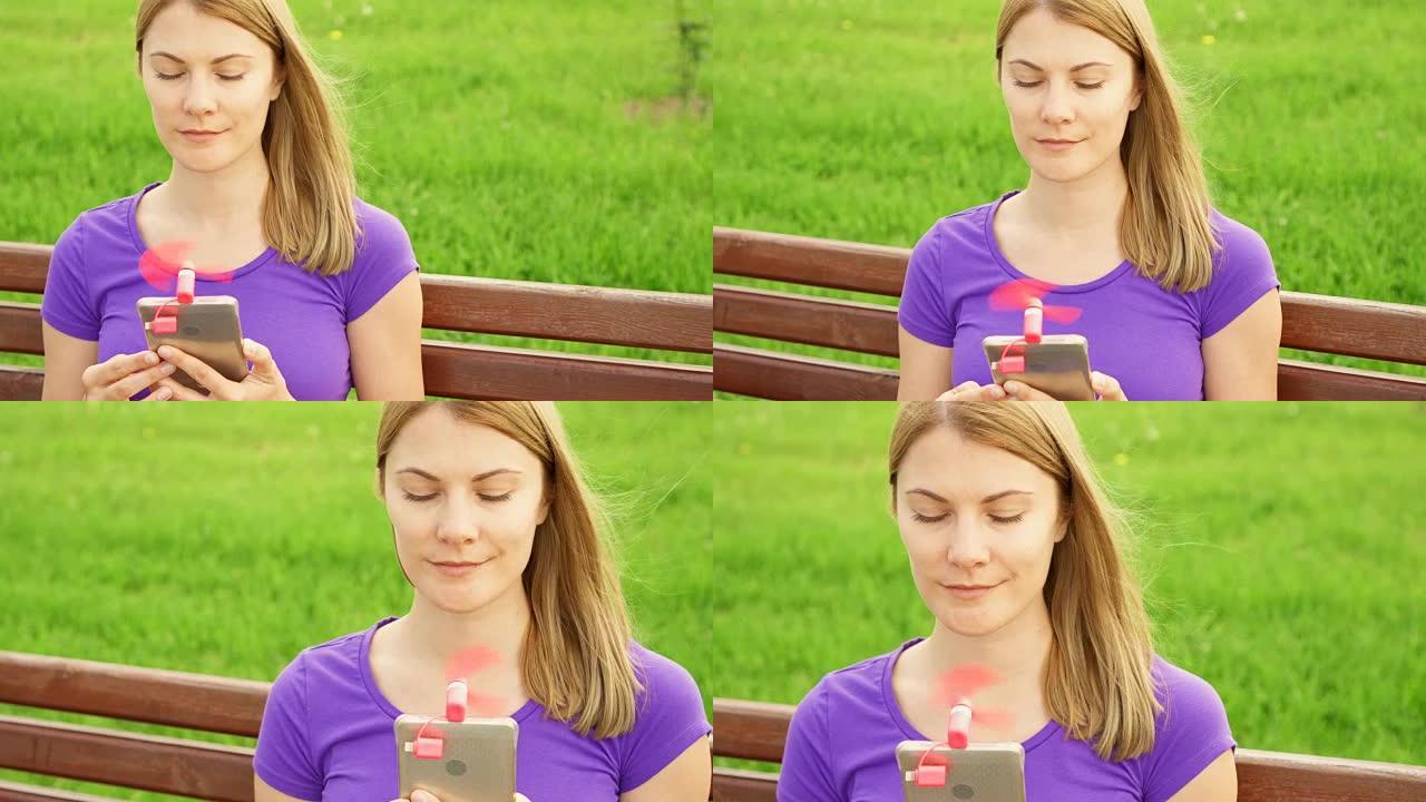 女人在炎热的时候在公园里用手机上的便携式呼吸机。用小风扇冷却