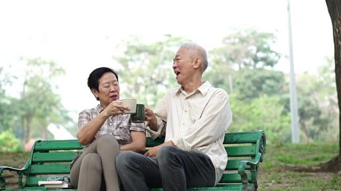 慢动作快乐的亚洲老年夫妇在早间公园坐着喝茶、咖啡。交谈和表达爱