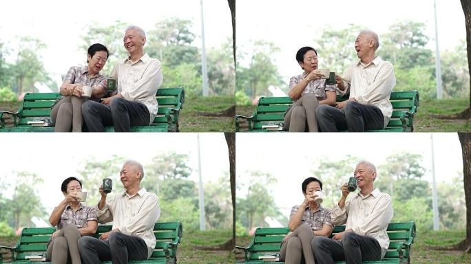 慢动作快乐的亚洲老年夫妇在早间公园坐着喝茶、咖啡。交谈和表达爱