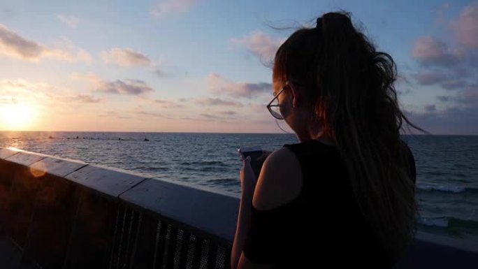 带智能手机的年轻女孩在特拉维夫的地中海上抵御美丽的日落。