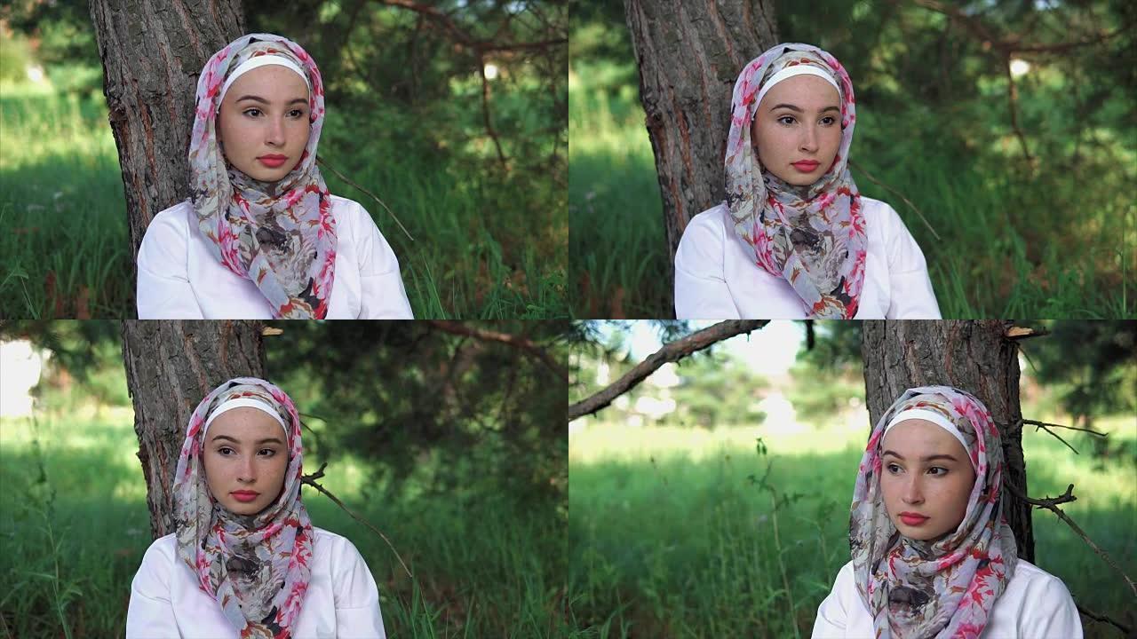 一名戴着穆斯林头巾的妇女坐在公园一棵树旁的草地上
