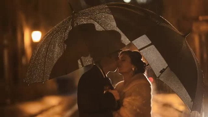 美丽的黑色风格的情侣在夜晚的城市街道上在雨中接吻，在雨伞下躲避雨