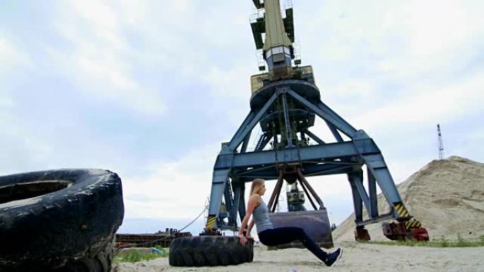 穿着灰色上衣和黑色绑腿的运动女子使用大型拖拉机轮进行力量锻炼，并进行俯卧撑训练手臂。在海滩上，在货港