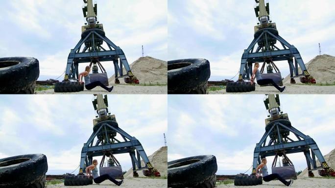 穿着灰色上衣和黑色绑腿的运动女子使用大型拖拉机轮进行力量锻炼，并进行俯卧撑训练手臂。在海滩上，在货港