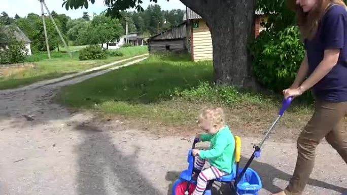 年轻女子母亲在小三轮车上推着金发碧眼的蹒跚学步的女孩。