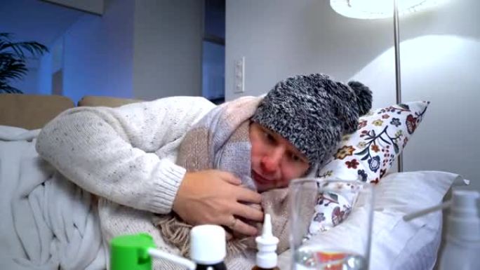 生病的男人戴着温暖的帽子和围巾在床上咳嗽并测量温度