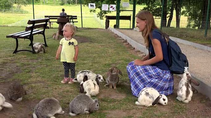 开朗的母亲和蹒跚学步的女儿在动物园里的一群兔子之间