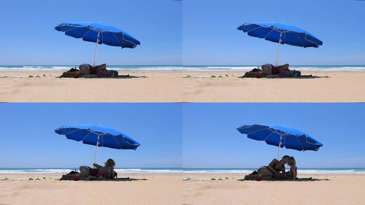 婴儿和母亲躺在海滩上的蓝色阳伞上