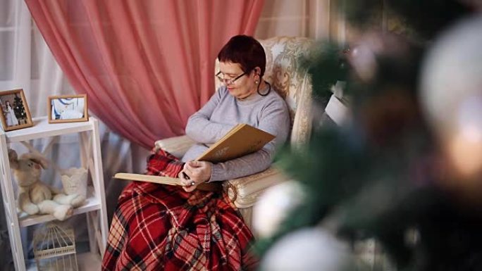一位戴着眼镜的老妇坐在火炉旁的扶手椅上，翻阅着带有照片的相册。除夕。