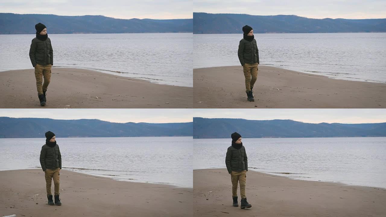 穿着夹克，黑帽子和脖子上的围巾的男孩在海滩上行走。在一个视图的背景下。美丽的大海，迷人的山脉，低波