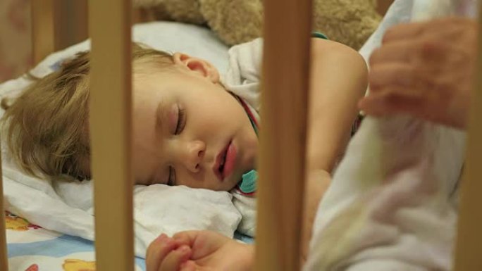 可爱的婴儿睡在婴儿床里。母亲盖好毯子