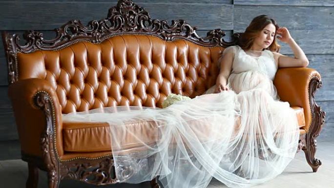 穿着豪华礼服的怀孕年轻新娘坐在皮沙发上