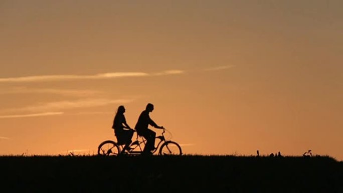 美丽的夫妇在夕阳下骑双人自行车的剪影。夏季自然背景与美丽的云在天空近距离