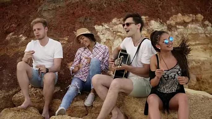一群年轻的时髦朋友坐在海边的岩石上，弹吉他，唱歌和跳舞。慢动作镜头