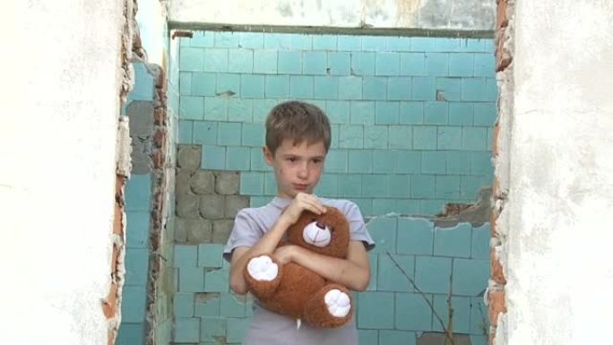 肮脏的小孤儿男孩站在一个柔软的玩具上，在被毁房屋废墟的背景下拍拍她