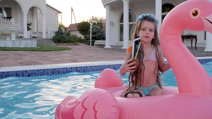 可爱的小女孩在游泳池里漂浮着一个空气吻，孩子坐在充气火烈鸟上喝甜饮料，丰富的周末