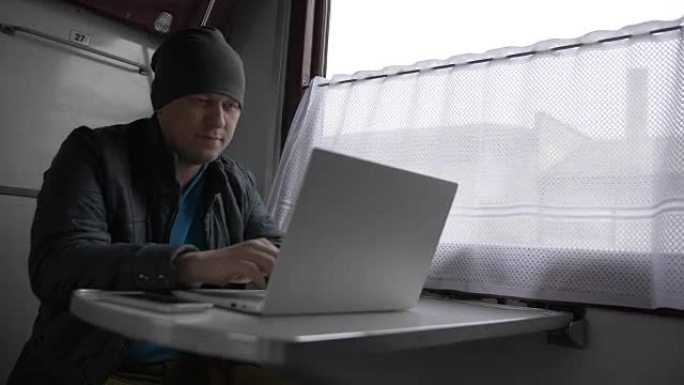 火车上的一个年轻人，在笔记本电脑上打印，微笑着，放下笔记本电脑的盖子，直视他的面前