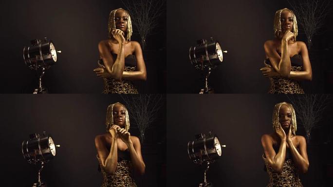 创意超现实的肖像性感非洲裔美国女模特与光泽的金色化妆和头戴相机摆姿势。青铜车身漆，黑色工作室背景。时