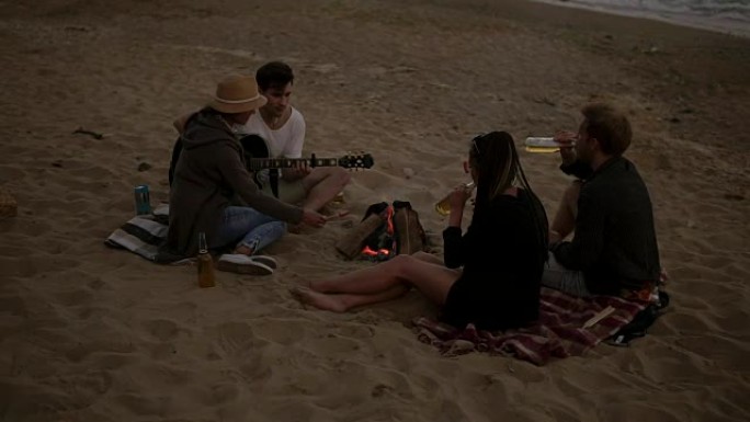 年轻的朋友坐在海边的火炉旁，烤香肠，喝啤酒，弹吉他。慢动作镜头