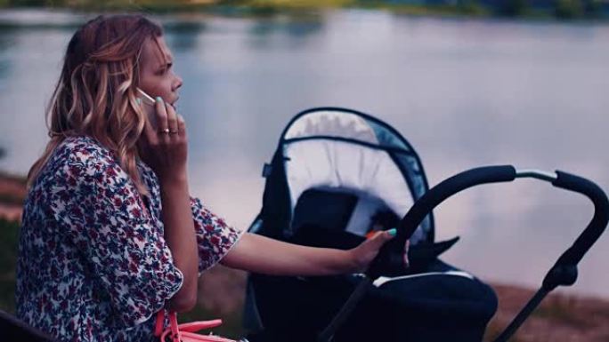 母亲摇摆婴儿车，在湖边的夏季公园打电话。阳光明媚