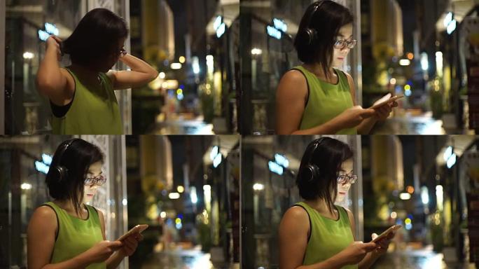 戴着耳机的亚洲女人晚上站在街上听手机音乐。