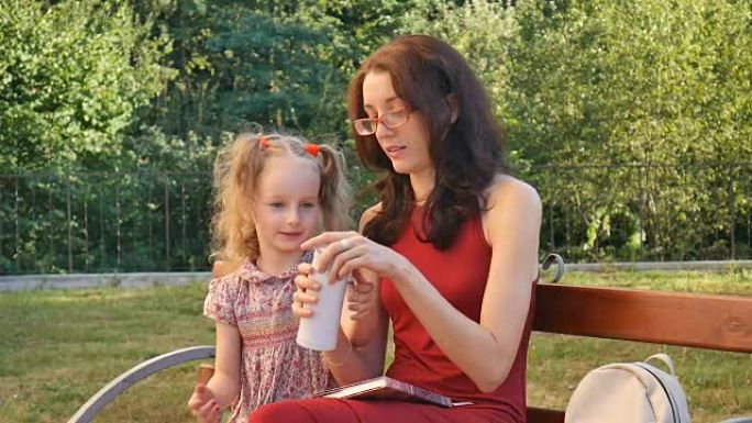 年轻的母亲戴着眼镜阅读，小女儿坐在公园的长凳上。两个马尾辫的女孩从瓶子里喝饮料。