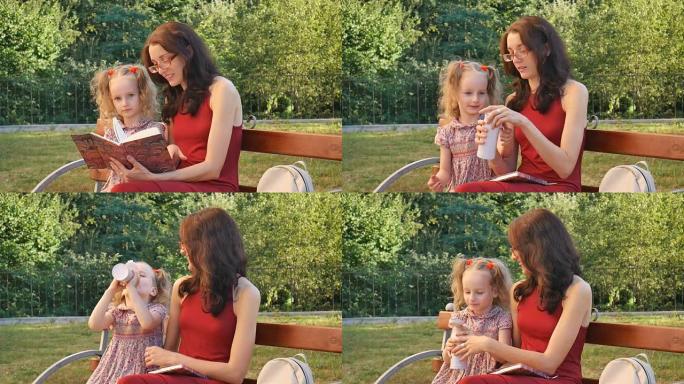 年轻的母亲戴着眼镜阅读，小女儿坐在公园的长凳上。两个马尾辫的女孩从瓶子里喝饮料。