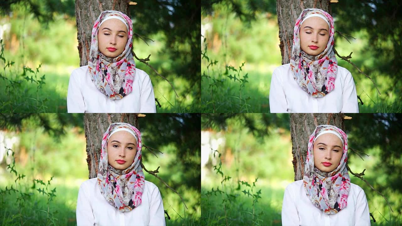 一位沉思的年轻穆斯林妇女的肖像，她坐在一棵树旁的森林中
