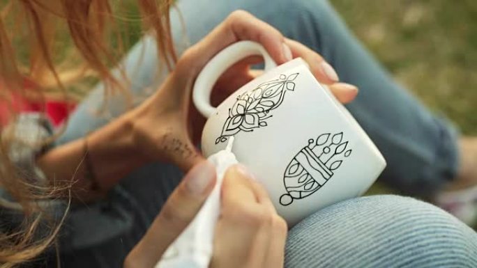 女性双手在白色杯子上画图案的特写镜头。晚上女孩坐在草地上画装饰品。