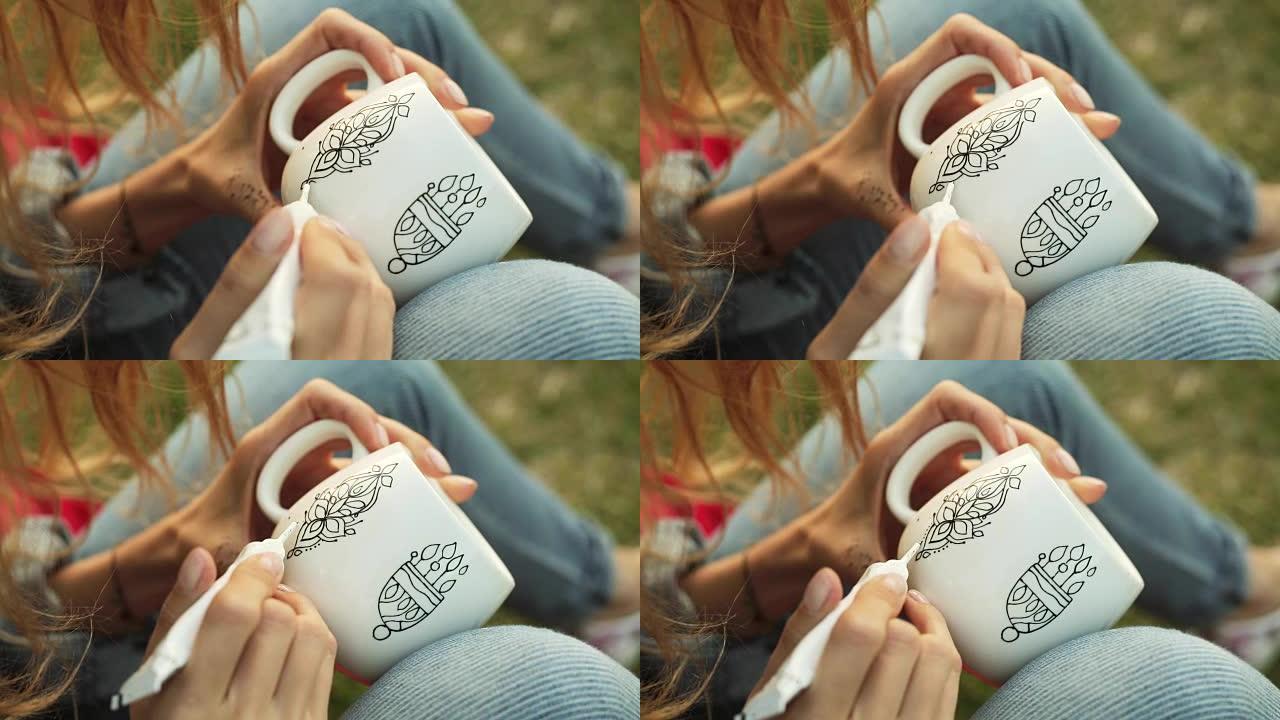女性双手在白色杯子上画图案的特写镜头。晚上女孩坐在草地上画装饰品。