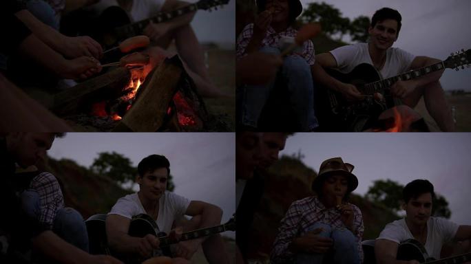 海滩火炉上烤香肠的特写视图。晚上，一群年轻开朗的人坐在海边的火炉旁，烤香肠和弹吉他。慢动作镜头