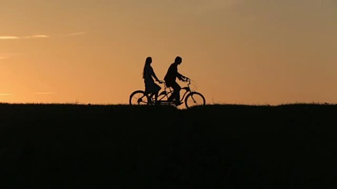 美丽的夫妇骑双人自行车而日落剪影。夏季自然背景与美丽的云在天空近距离
