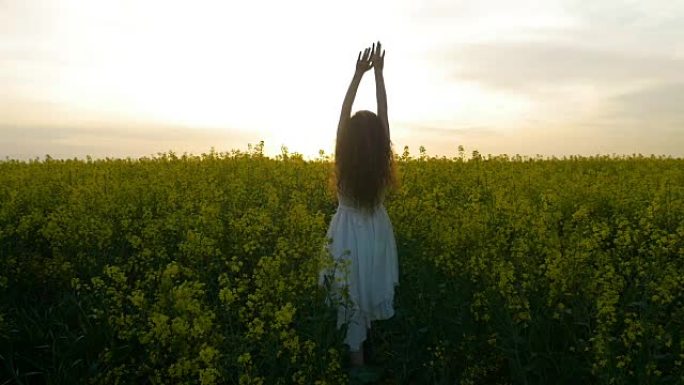 年轻的乡村女孩穿着白色的衣服，在高大的油菜籽花丛中举起双臂，感到和平