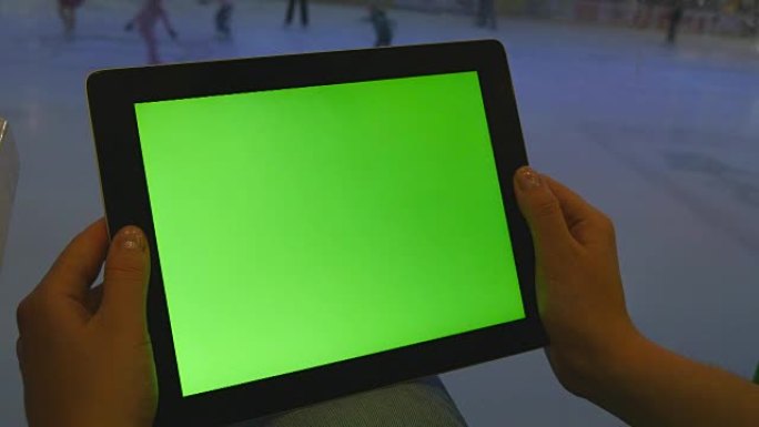 在购物中心使用绿屏平板电脑的女性双手。很多人在后台的室内溜冰场上滑冰
